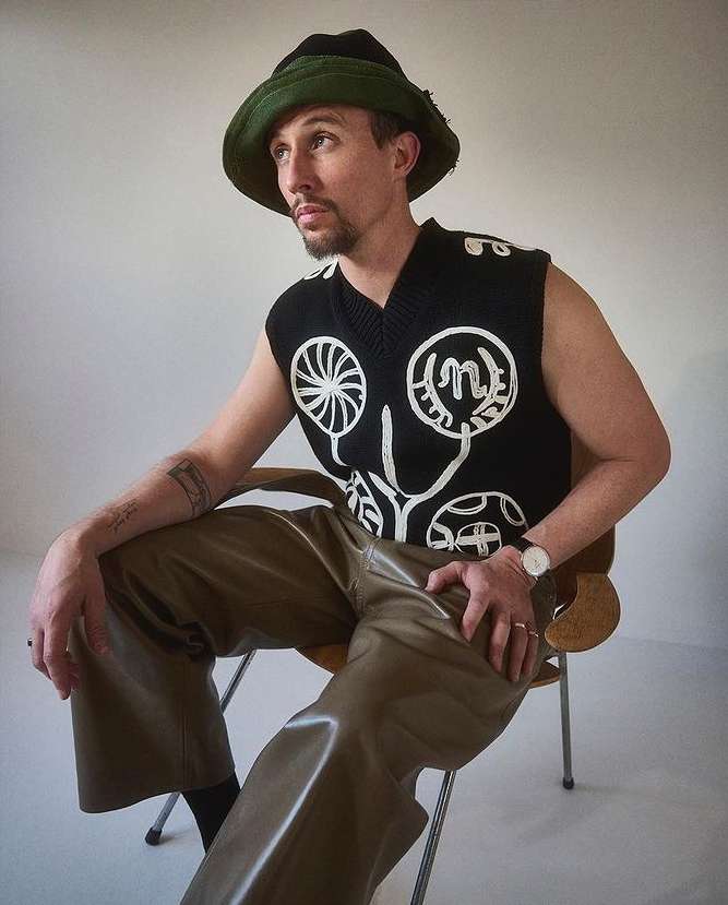 An adult male sits in a chair, wearing a black sweater vest with white graphics, olive green pants, and a dark green bucket hat.