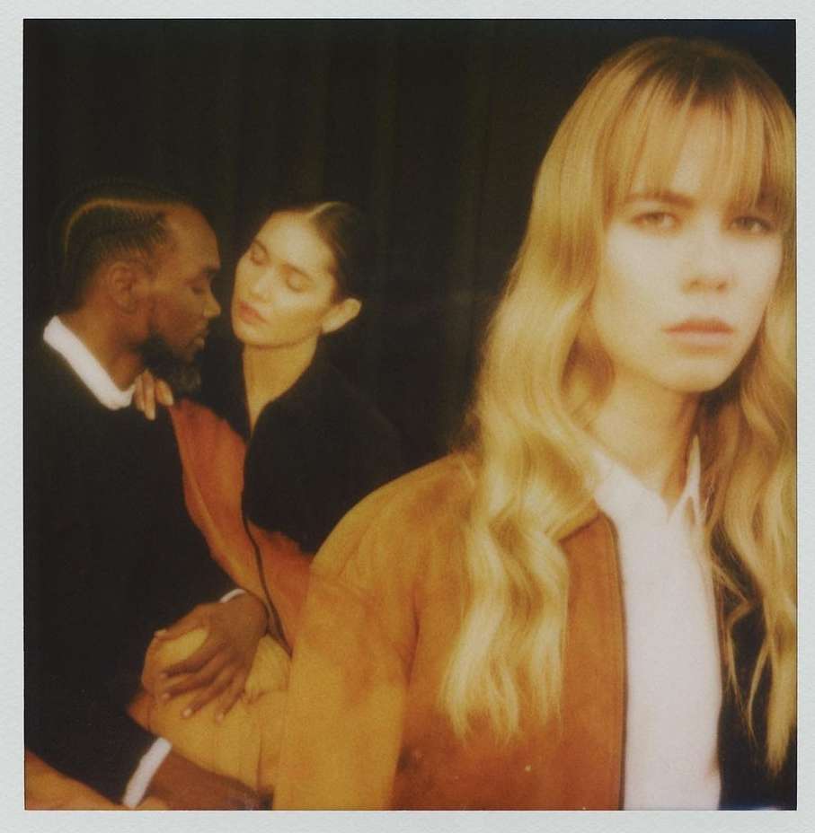 Three people: a blonde woman in the foreground, a woman behind her, and a man embracing her on the left side of the frame.