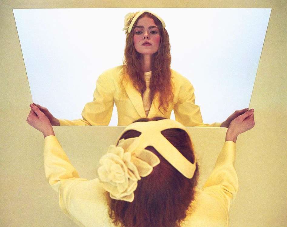 A woman in yellow jacket looks in mirror, reflecting an image of a woman wearing a flower-decorated headpiece. Studio setting, soft lighting.