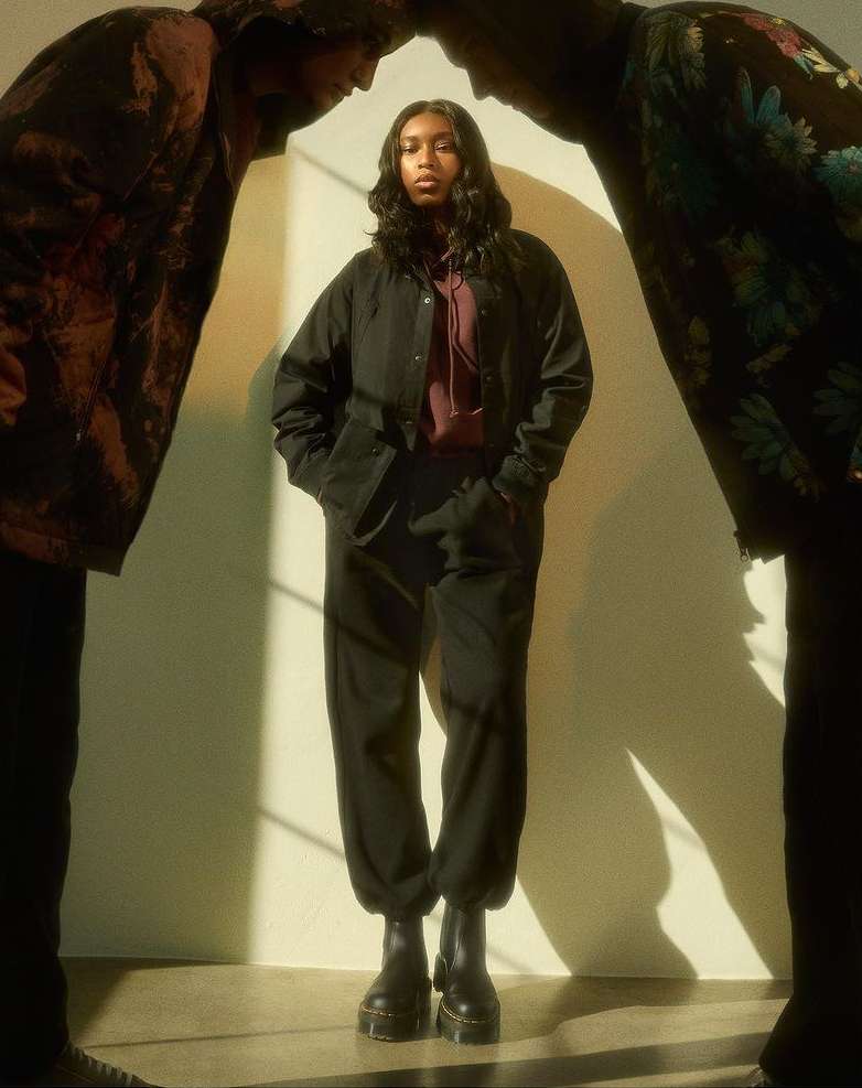 A woman wearing black jacket and pants is standing between two men in dark outfits in a studio, with soft lighting creating shadows.