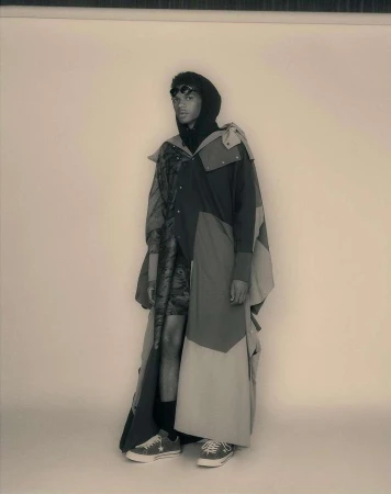 Full-length portrait of a male model wearing a dark hooded sweatshirt, a long panelled coat, and sneakers against a seamless backdrop