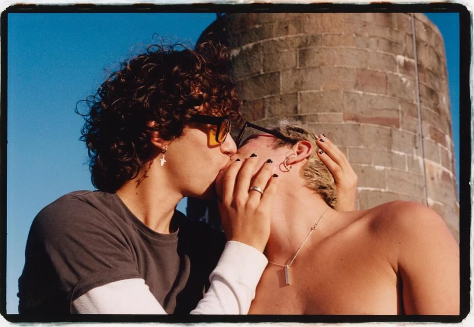 A couple kisses in a close-up shot in front of a brick tower against a bright blue sky. One man has curly hair and sunglasses.