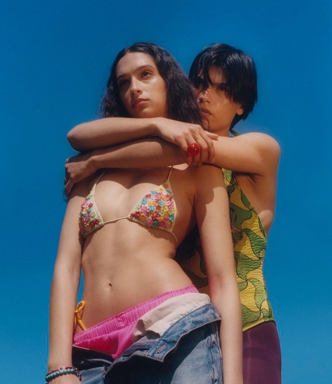 Two women pose outdoors under a bright blue sky. One woman wears a floral bikini top and jeans, while the other wears a yellow top.