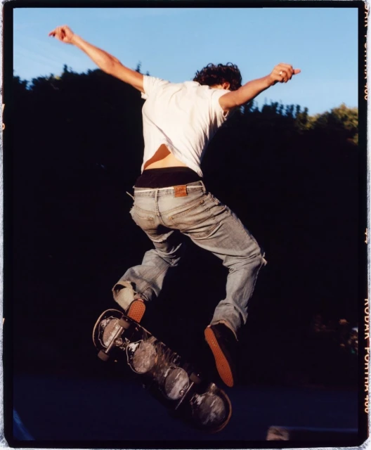 A person does a skateboarding trick. The skateboarder has a white t-shirt and denim jeans on. A background of trees and a blue sky.