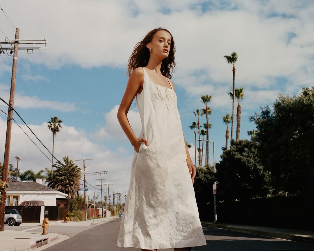 Woman in a white dress stands on a street with palm trees under a blue sky with clouds, creating a sunny outdoor fashion portrait.