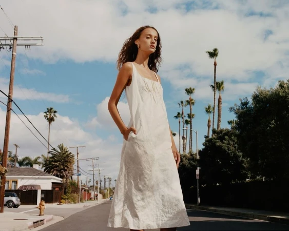 Woman in a white dress stands on a street with palm trees under a blue sky with clouds, creating a sunny outdoor fashion portrait.