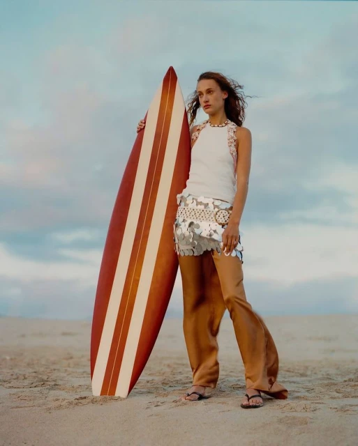 Adult female model stands on a beach holding a surfboard with tan and white stripes against a cloudy sky background during the day.