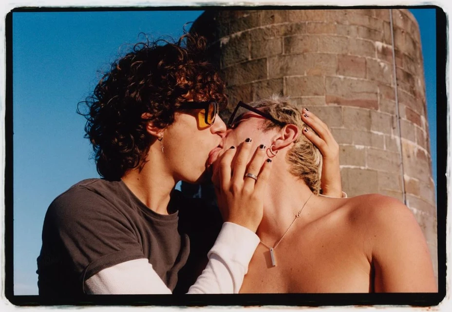 Two young adult males share a kiss in an outdoor setting with a stone tower visible in the background under a clear blue sky.