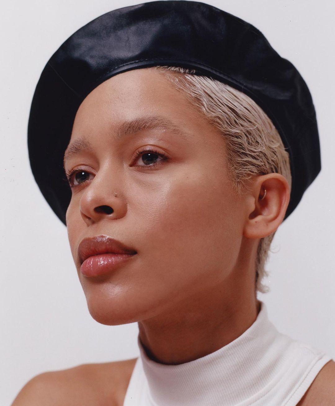 Close-up portrait of a Black female adult with short blonde hair wearing a black beret and a white turtleneck sweater on a neutral background.