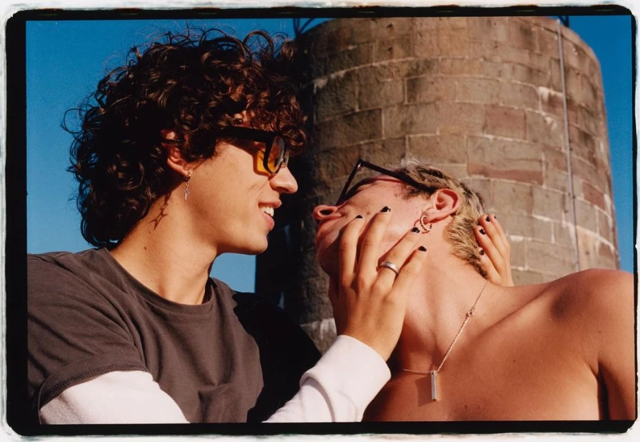 A curly-haired man smiles at another man with platinum-blond hair being touched on the face in front of a brick building.