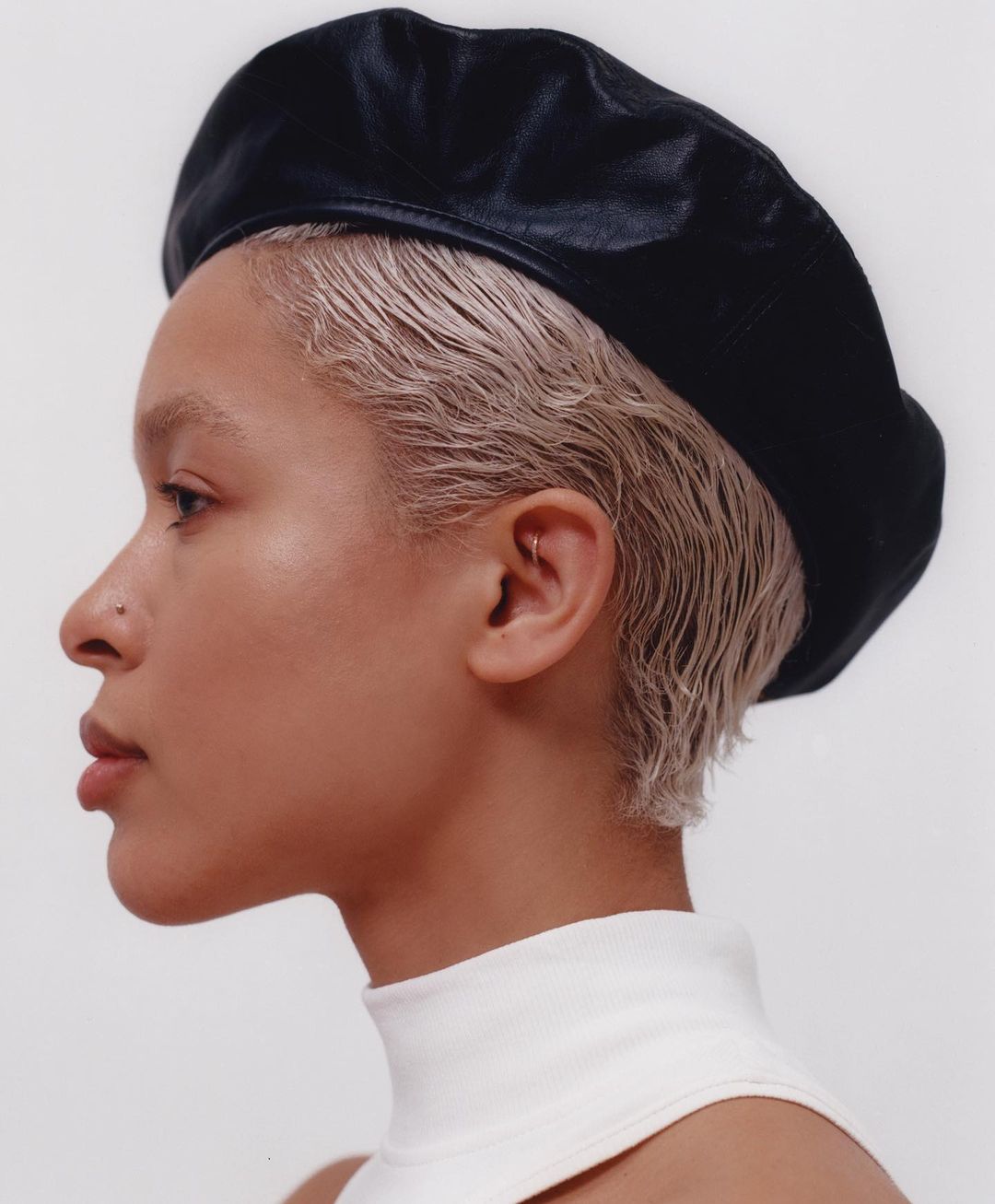 Profile portrait of a young adult woman with platinum blonde hair wearing a black beret against a clean, bright white background.