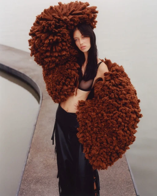 Adult female fashion model poses in front of water wearing black skirt, sheer top and large brown faux fur overcoat. 