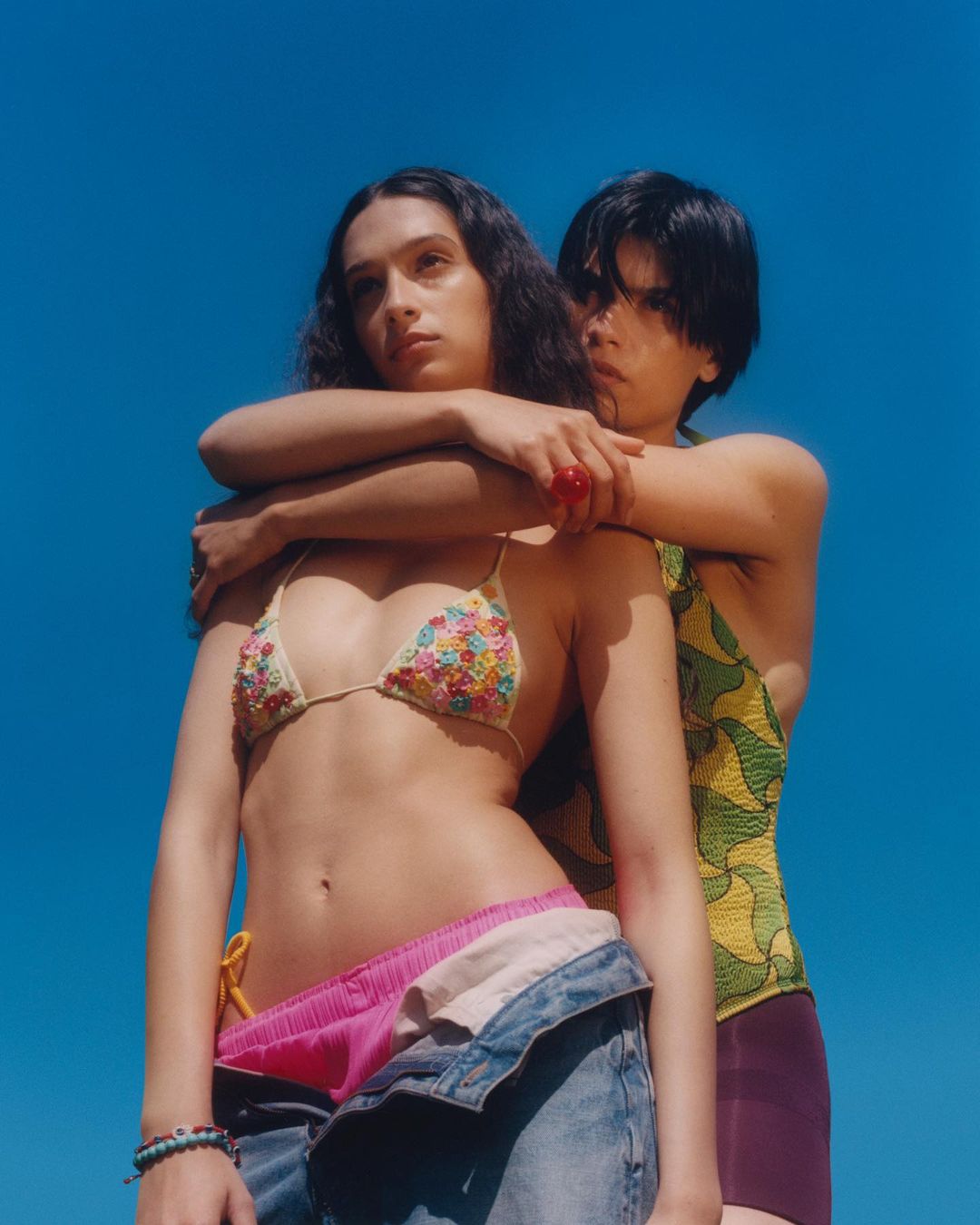 Two women pose together against a vibrant blue sky. One woman is wearing a floral bikini top and open denim pants, while the other is embracing her.