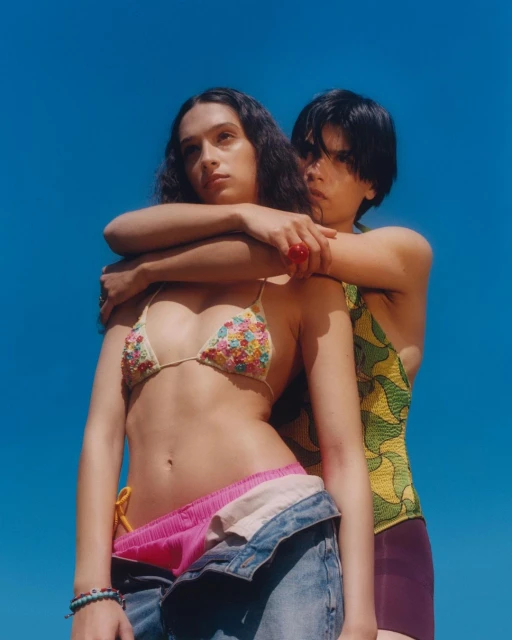 Two women pose together against a vibrant blue sky. One woman is wearing a floral bikini top and open denim pants, while the other is embracing her.