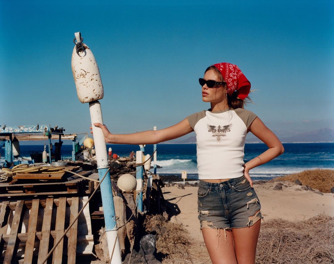 Adult female poses next to ocean buoy on a sunny day at the beach wearing a white tee shirt, red bandana, and blue denim shorts.