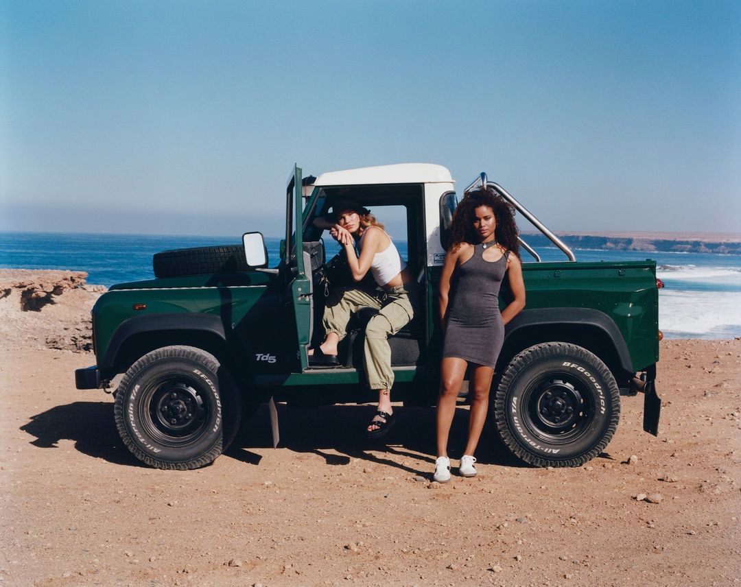 Two women pose with a vintage green Land Rover on a sandy beach with the ocean and a rocky coastline in the background under a clear blue sky.