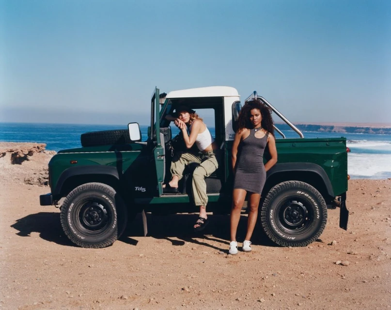 Two women pose with a vintage green Land Rover on a sandy beach with the ocean and a rocky coastline in the background under a clear blue sky.