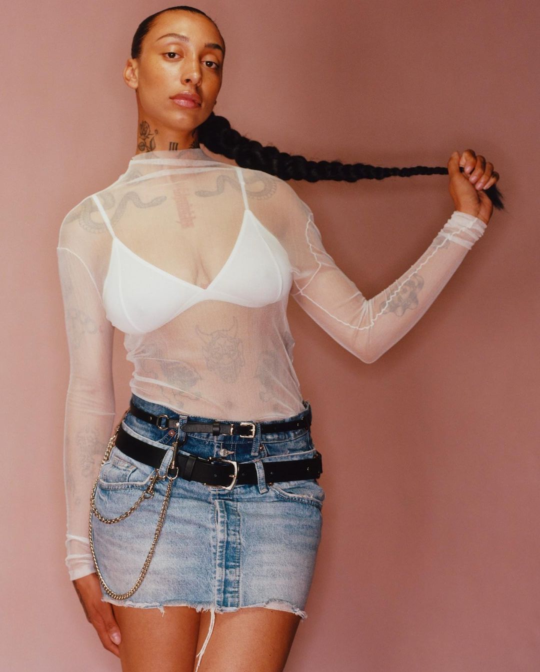 Portrait of a young adult female with a long braid, wearing a white top and denim skirt, posing against a pink backdrop in a studio.