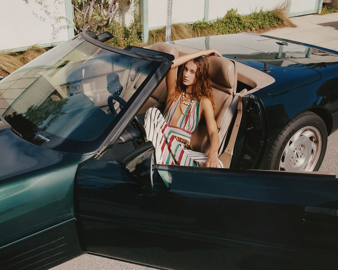A brunette woman in a colorful striped outfit sits in a dark green convertible car with tan leather seats, posing outside during daylight.