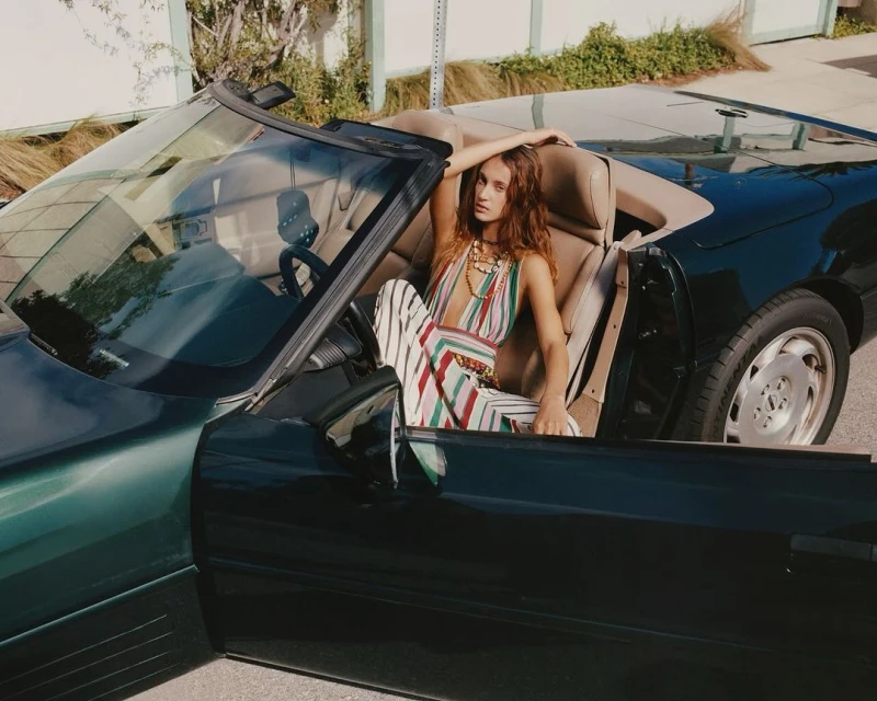 A brunette woman in a colorful striped outfit sits in a dark green convertible car with tan leather seats, posing outside during daylight.