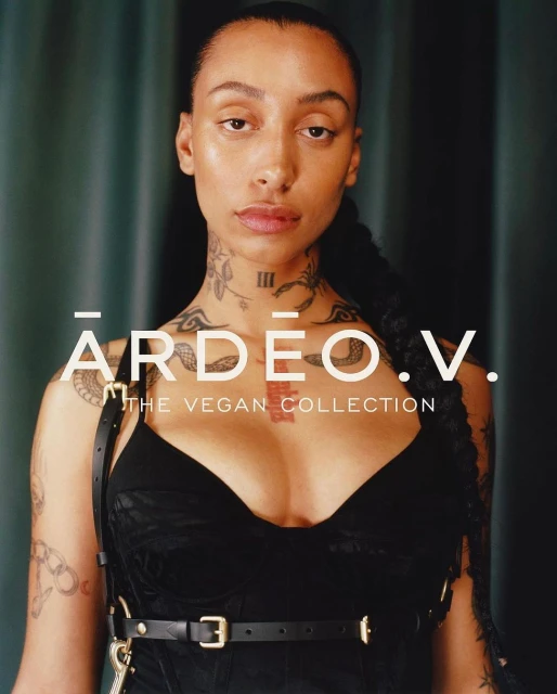 Close-up portrait of a woman with tattoos wearing a black top and harness against a dark green backdrop.