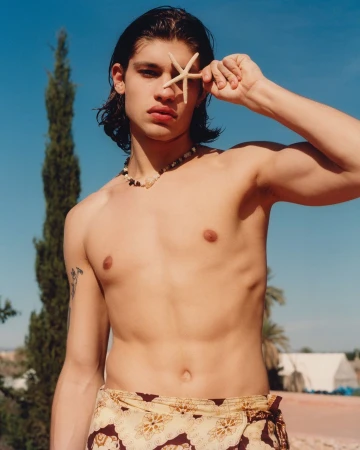 Shirtless young man poses outdoors with a starfish covering his eye, wearing a patterned wrap and necklace against a blue sky.