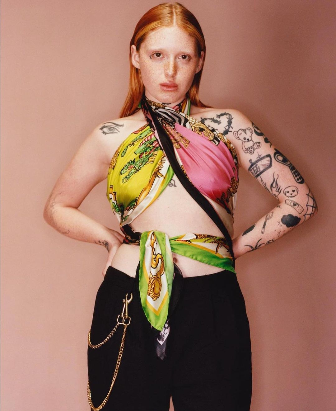 A red-haired person with tattoos poses in a studio, wearing a colorful scarf top and black pants with gold chain against a pink backdrop.