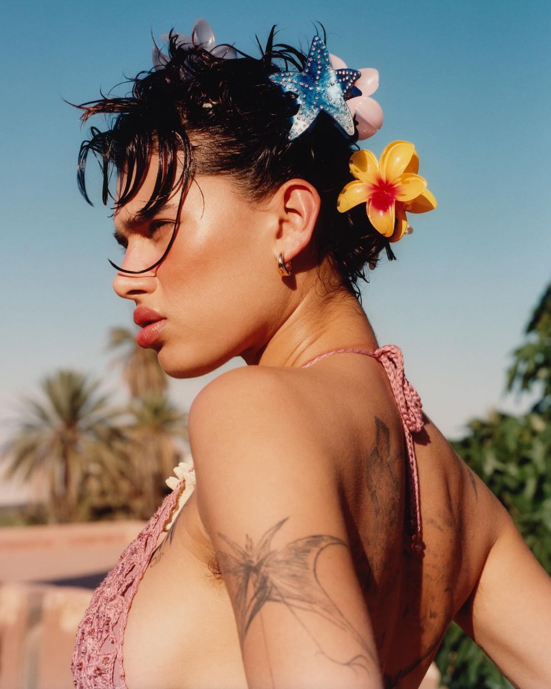 Profile portrait of woman with fair skin wearing a crochet top and decorative hair accessories against a blue sky and blurred background.