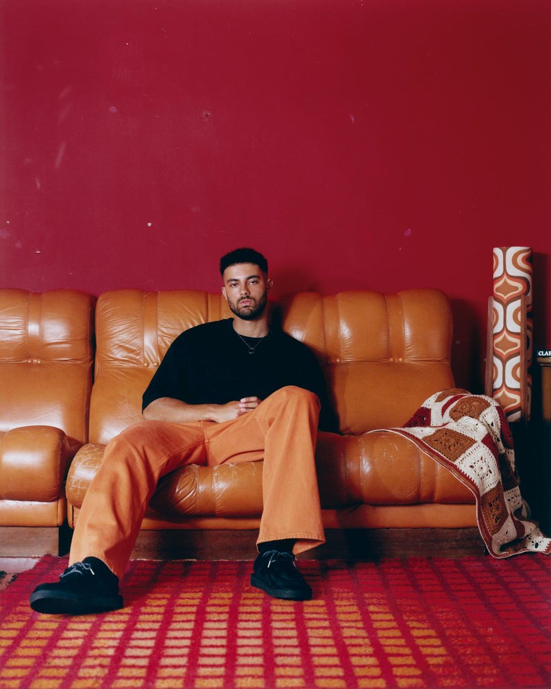 Man seated on an orange leather sofa wearing a black t-shirt and orange trousers against a dark red wall in London.