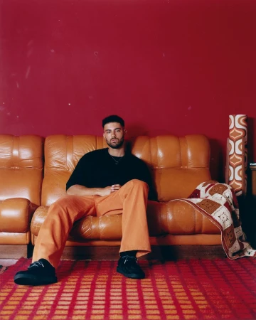Man seated on an orange leather sofa wearing a black t-shirt and orange trousers against a dark red wall in London.