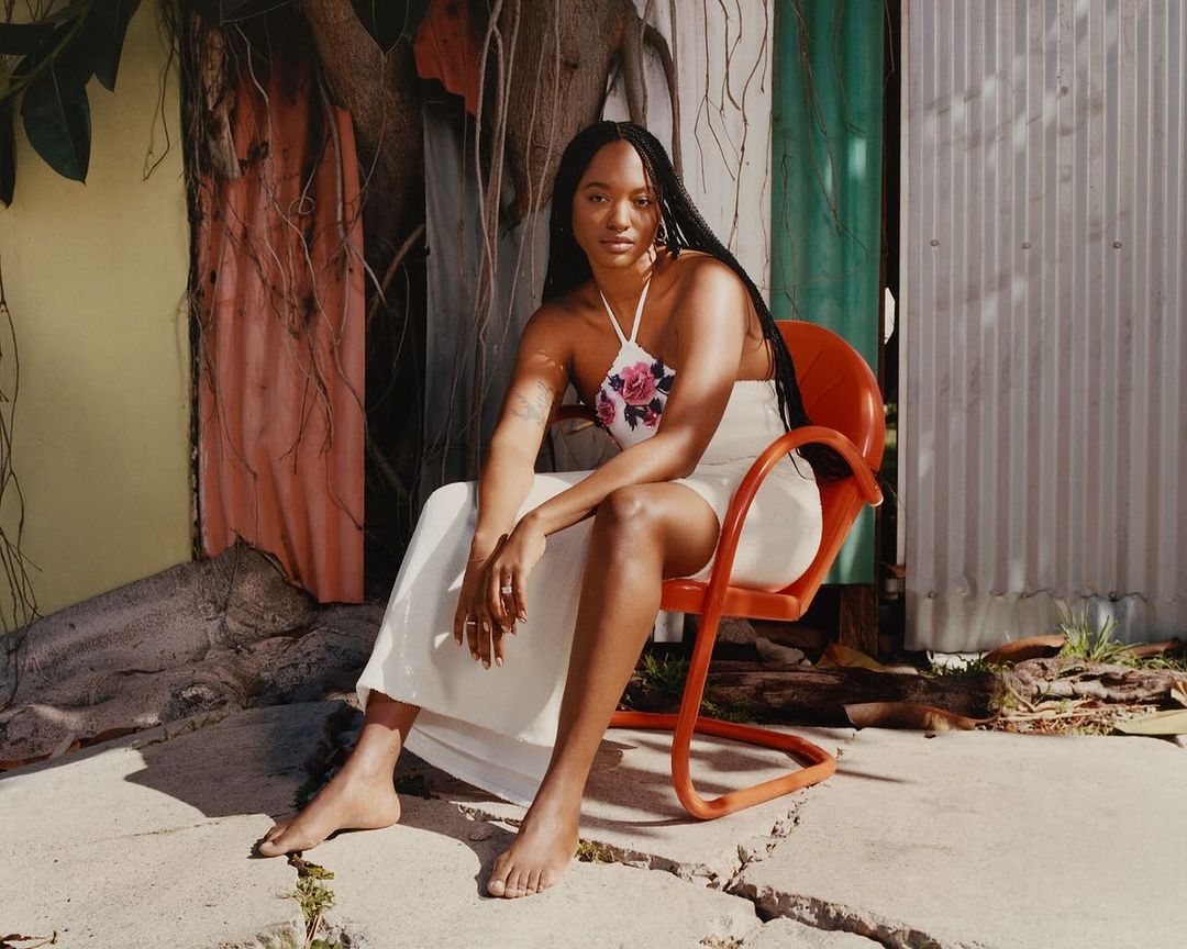 An adult woman with long dark hair sits in an orange chair wearing a white dress, with a corrugated metal wall in the background.