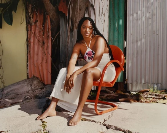 An adult woman with long dark hair sits in an orange chair wearing a white dress, with a corrugated metal wall in the background.