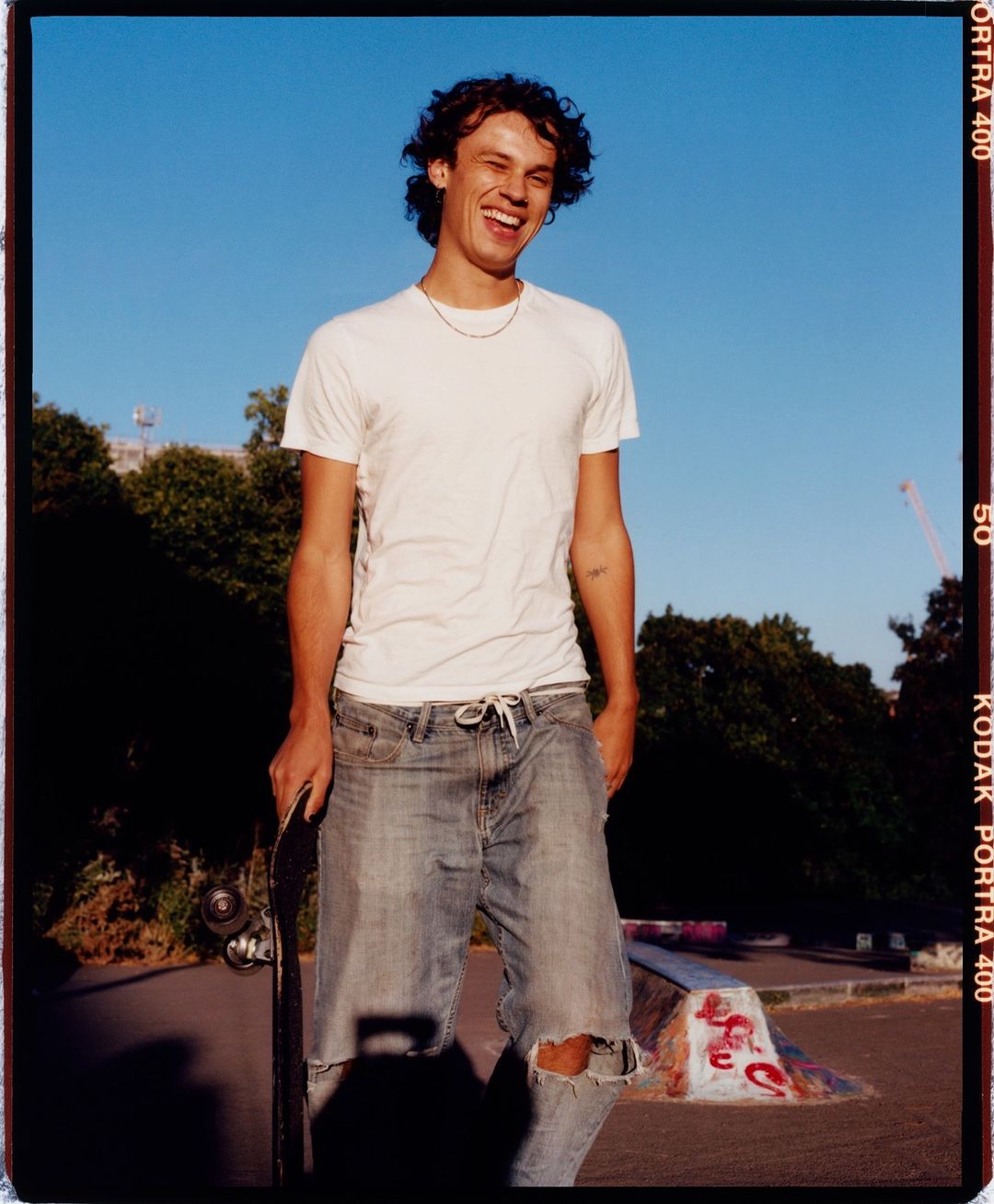 Smiling young man stands with a skateboard at a skate park on a sunny day, wearing a white t-shirt and ripped jeans.