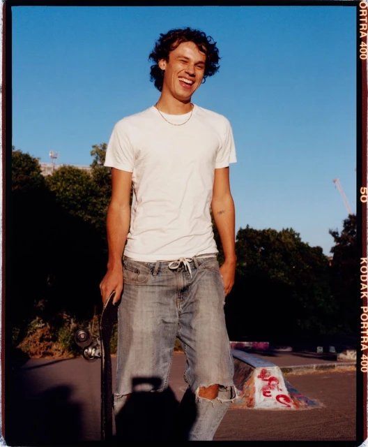 Smiling young man stands with a skateboard at a skate park on a sunny day, wearing a white t-shirt and ripped jeans.