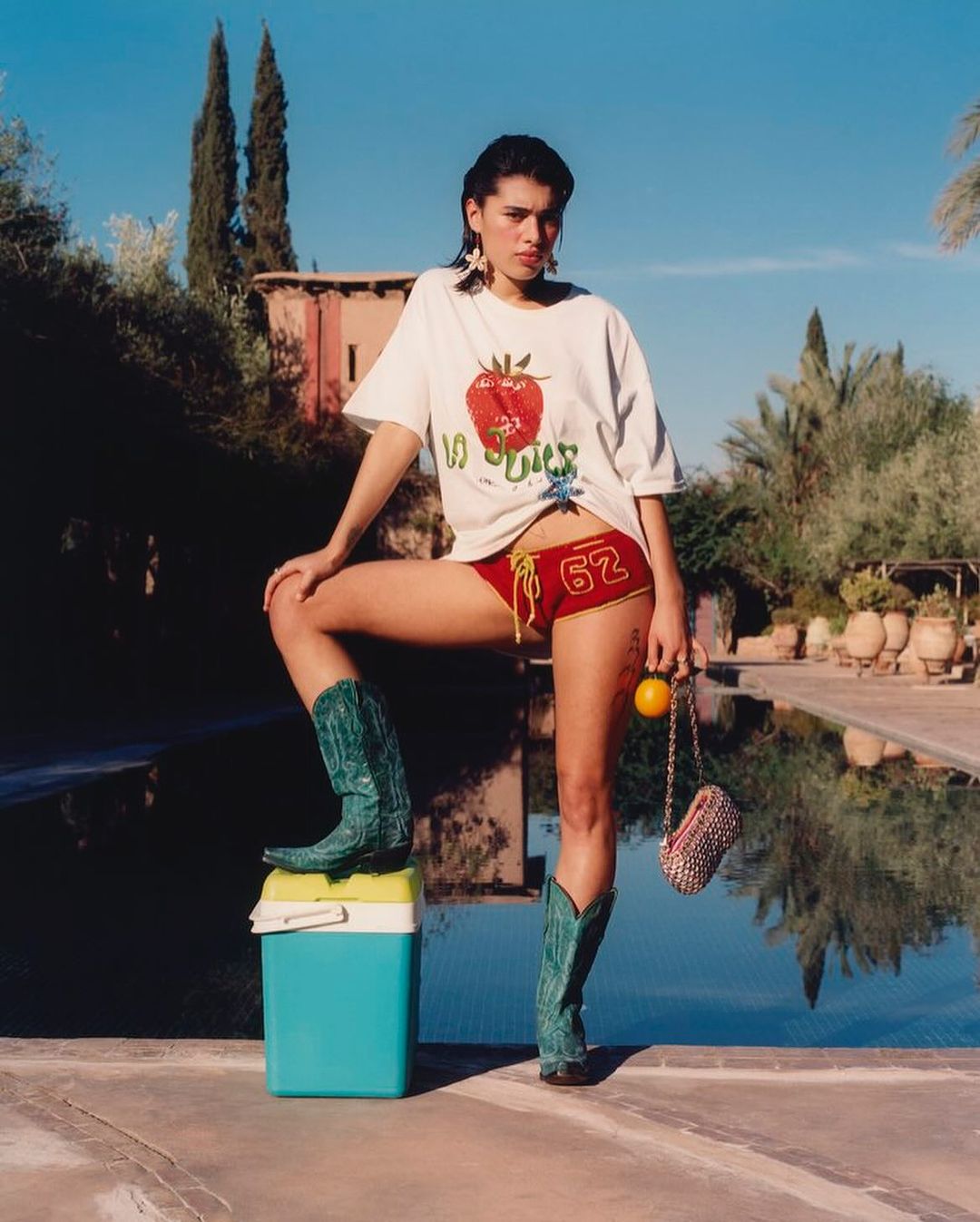 Young woman in T-shirt, red shorts and cowboy boots poses outdoors with a cooler, orange and purse in a pool-adjacent outdoor setting.