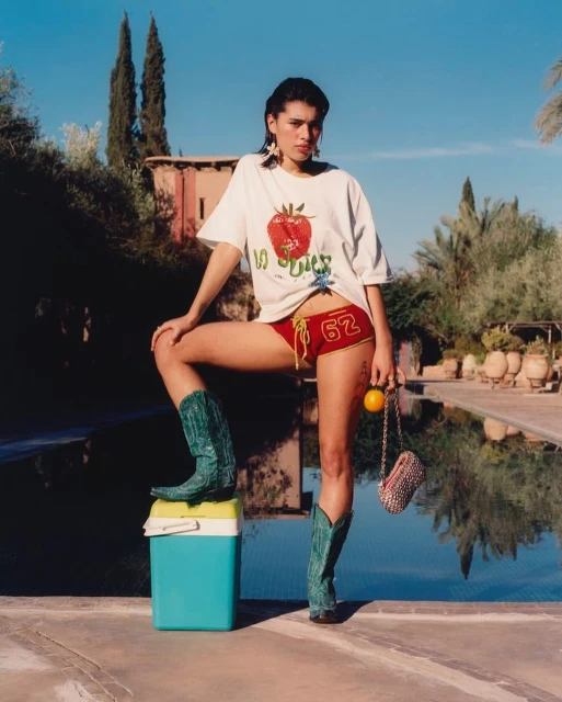 Young woman in T-shirt, red shorts and cowboy boots poses outdoors with a cooler, orange and purse in a pool-adjacent outdoor setting.
