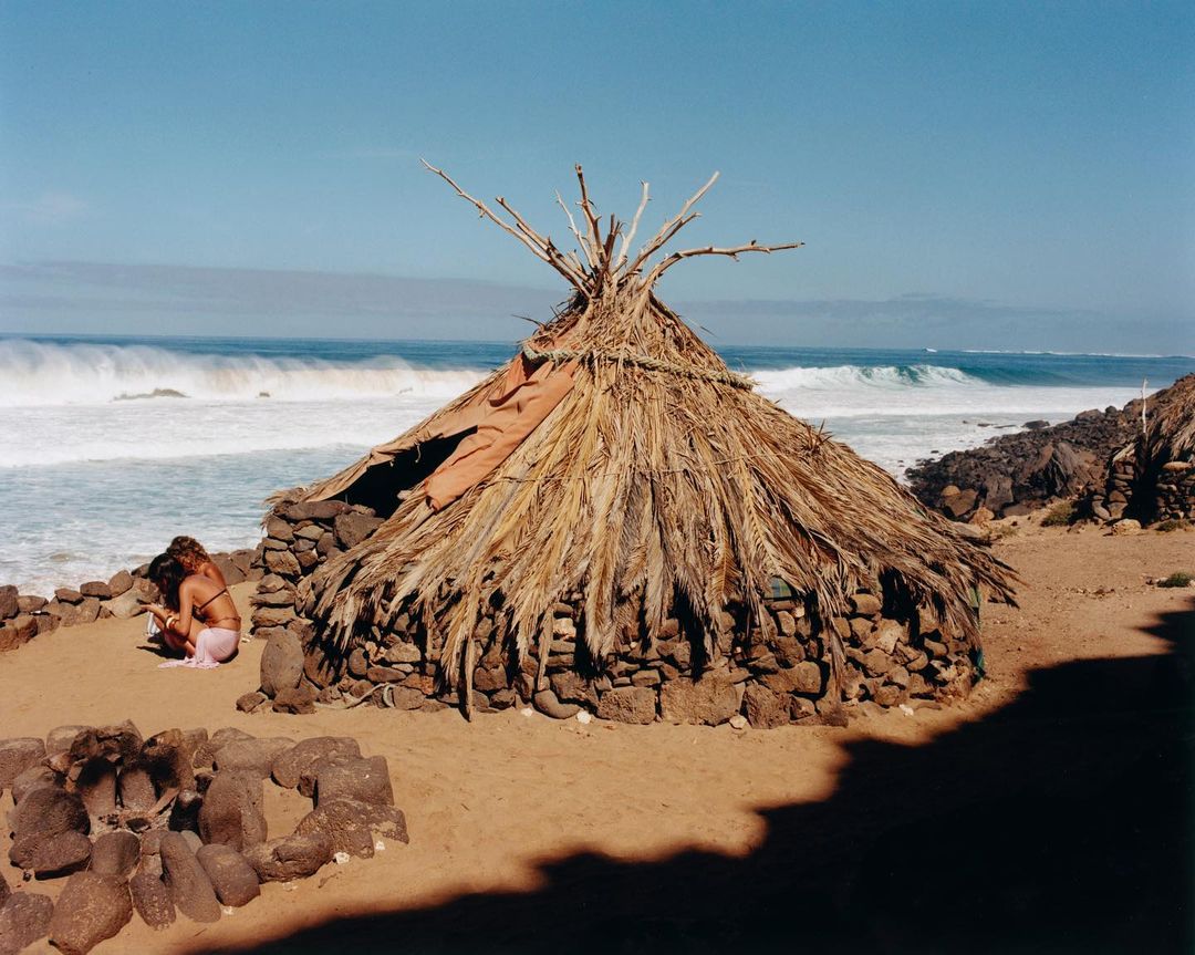 Two women sit near a hut with a thatched roof on a sandy beach. The ocean stretches to the horizon under a clear sky.