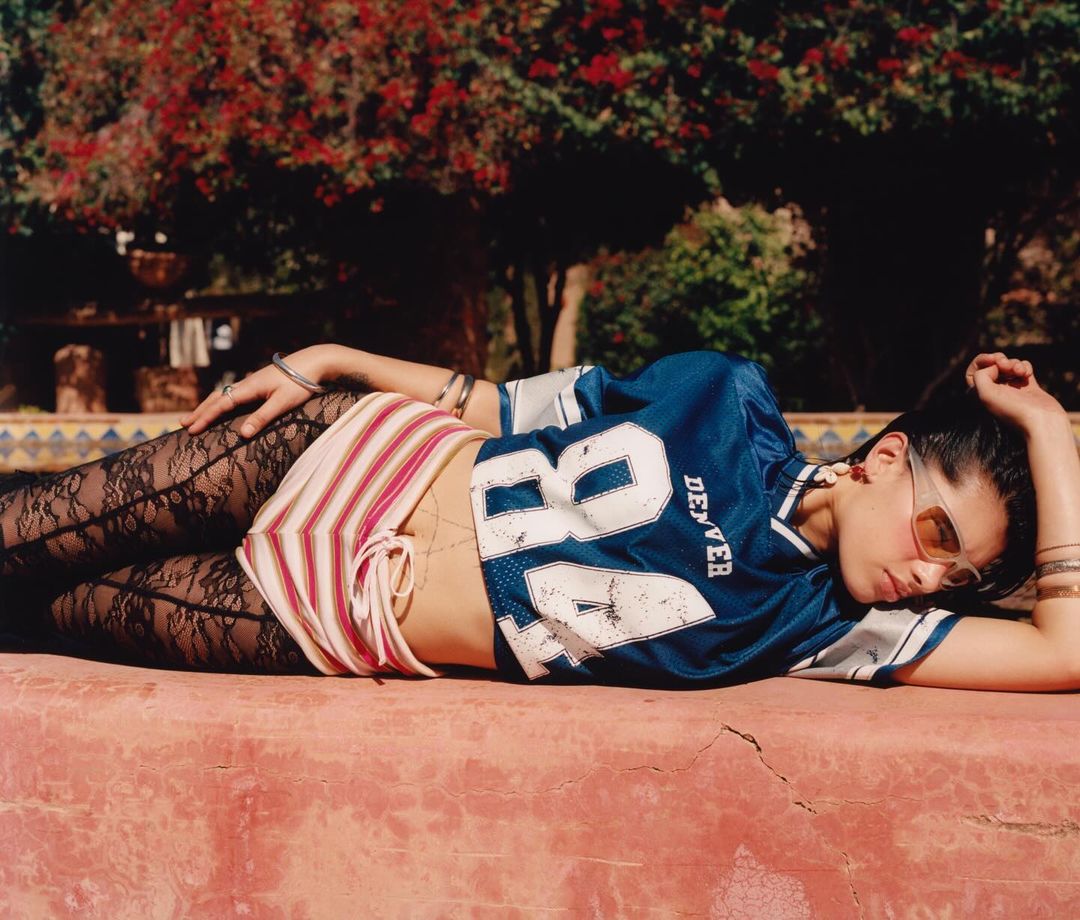 Young woman in football jersey lies on a red surface in a garden setting with trees, lace leggings, and sunglasses.