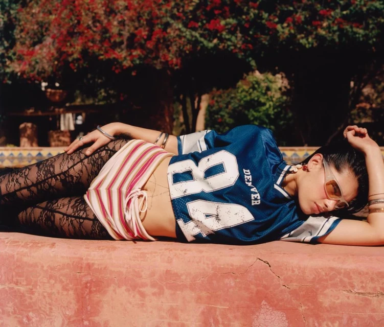 Young woman in football jersey lies on a red surface in a garden setting with trees, lace leggings, and sunglasses.