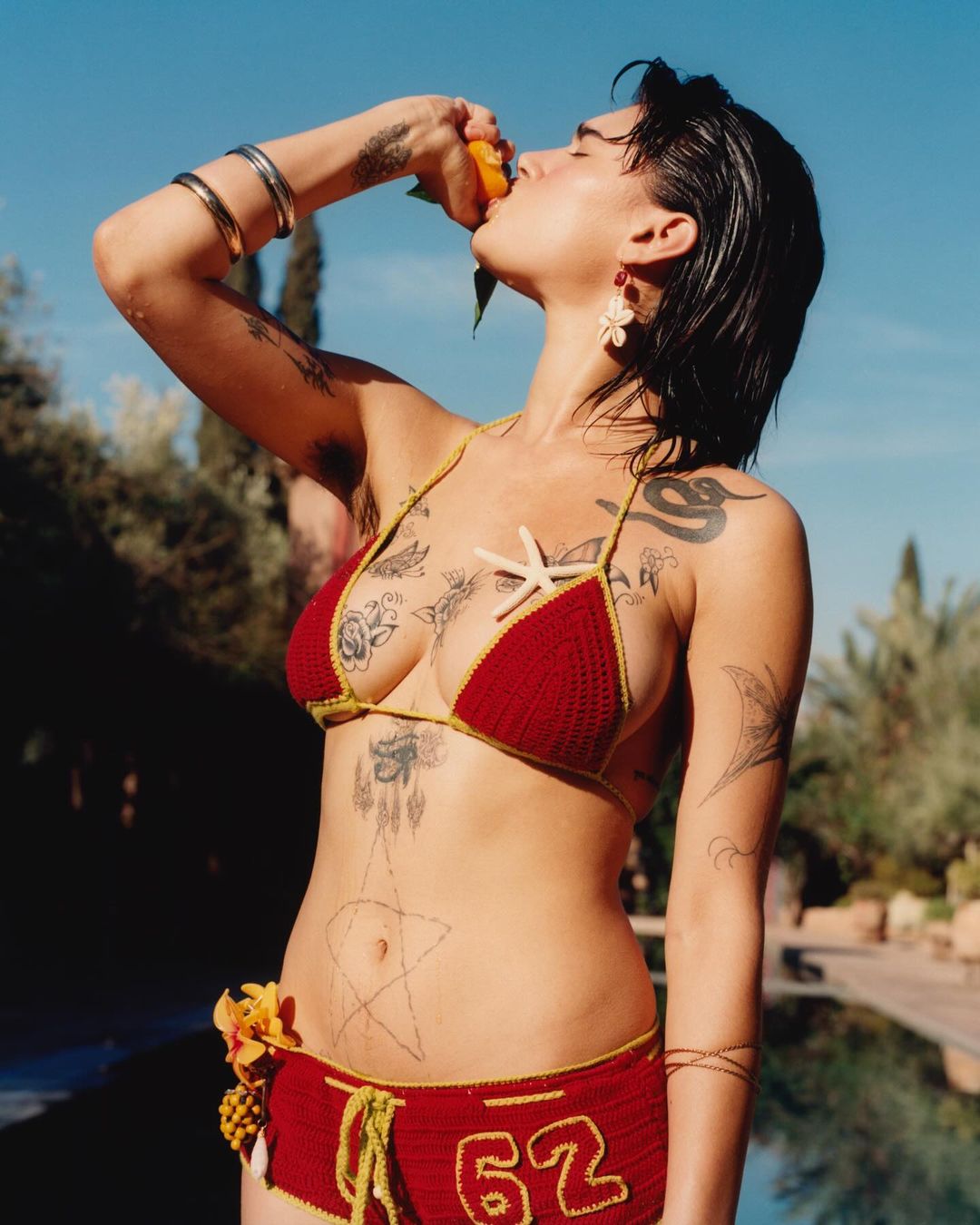Woman in red crochet bikini eating orange outdoors against blue sky with palm trees and swimming pool in background on sunny day.
