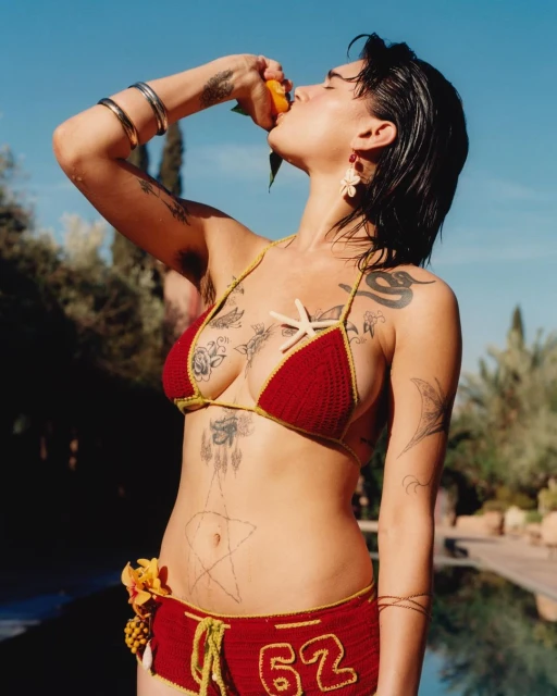 Woman in red crochet bikini eating orange outdoors against blue sky with palm trees and swimming pool in background on sunny day.
