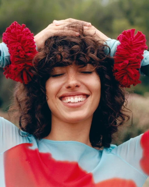 Smiling woman with brown curly hair and a blue and red patterned shirt, posing with hands on head with red flower-like wristbands.