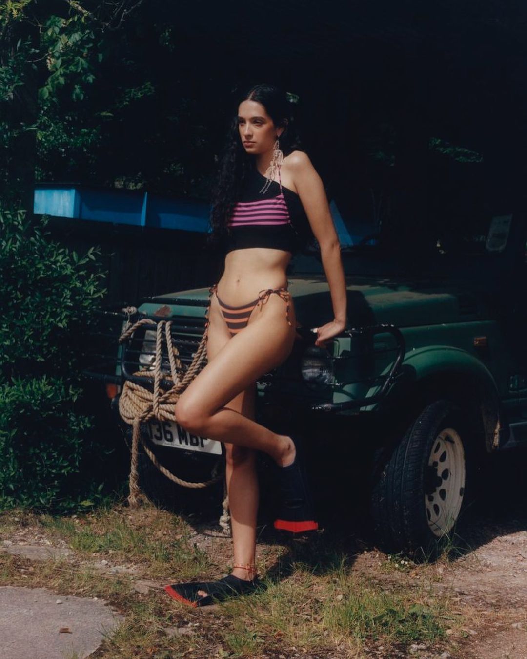 A woman in a bikini poses in front of a green vehicle outdoors, with a dark background of foliage and a dirt and grass ground.
