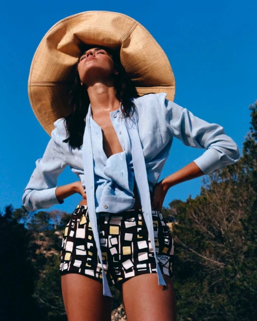 A woman wearing a large straw hat and shorts with a geometric pattern poses against a bright blue sky with trees in the background.