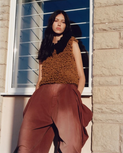 A young adult woman poses in a sleeveless top and wide-leg pants, set against a stone wall and window. Natural light illuminates the scene.
