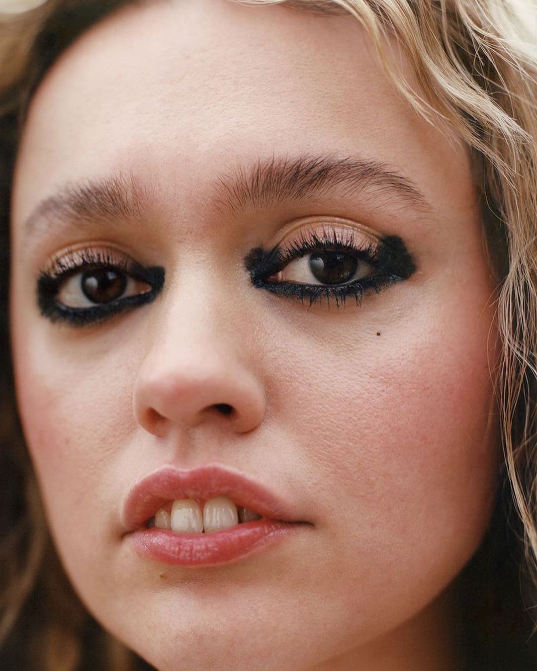 Close-up portrait of a young woman with heavy dark smudged eye makeup and wavy blonde hair.