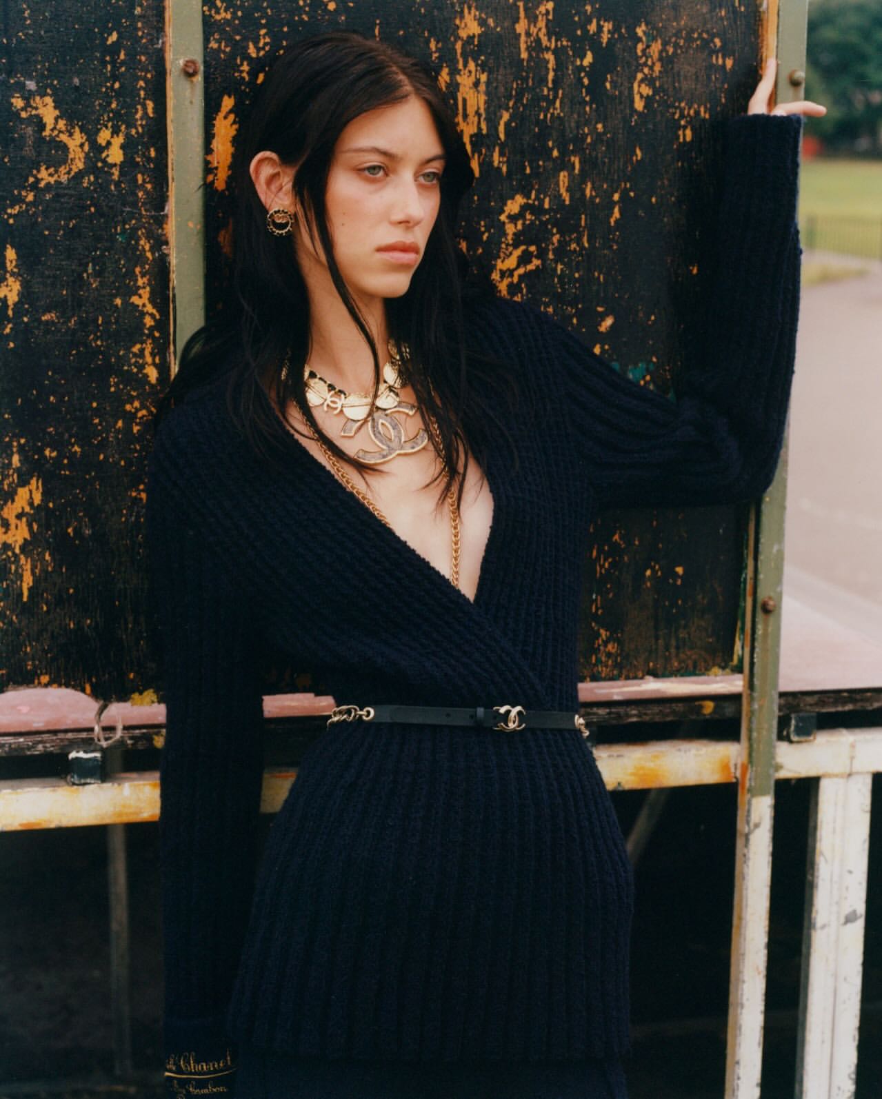 A woman with long dark hair poses against an aged panel wearing a black wrap sweater, black skirt, and gold Chanel jewelry.