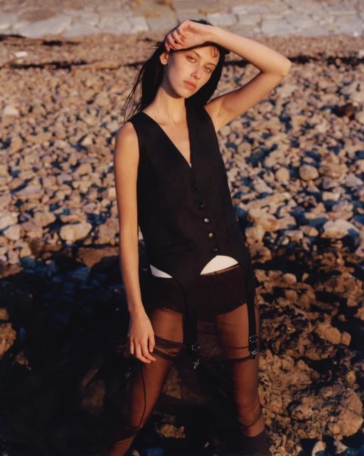 Portrait of a young woman in a black vest and sheer skirt posing on a rocky beach at golden hour, looking directly at the camera.