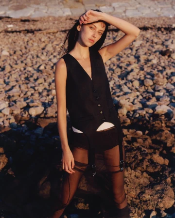 Portrait of a young woman in a black vest and sheer skirt posing on a rocky beach at golden hour, looking directly at the camera.
