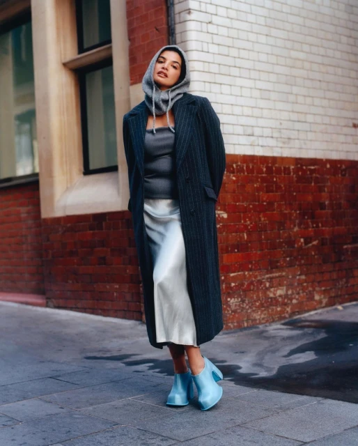 Woman poses in pinstripe coat and gray hoodie dress, accessorized with blue block-heel shoes, against a brick-and-stone wall backdrop.